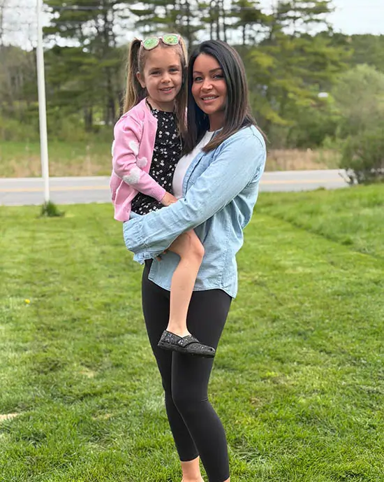 Assistant Manager, Stormy Wimber, standing on the grass holding her smiling young daughter in a pink sweater; trees and a road are visible in the background, highlighting moments of joy often cherished in families receiving specialized dementia care.